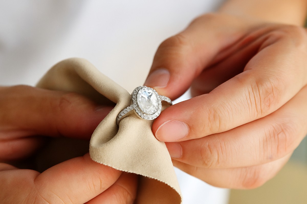 Une main nettoie délicatement une bague sertie de pierres avec un chiffon beige en microfibre, illustrant un geste doux d’entretien de bijoux en argent.